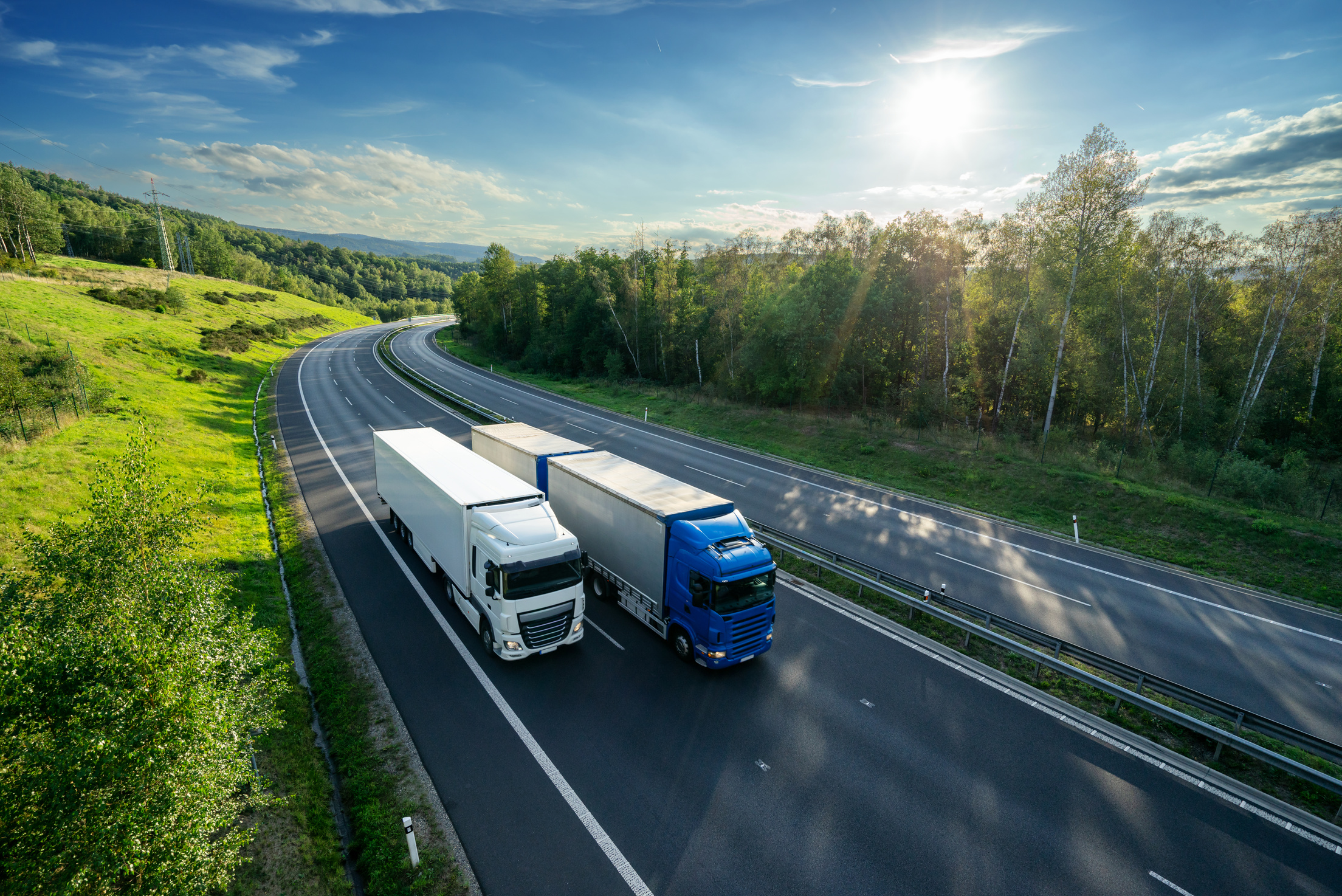 Blue truck overtaking white truck on an asphalt highway between forests under radiant sun. View from above.